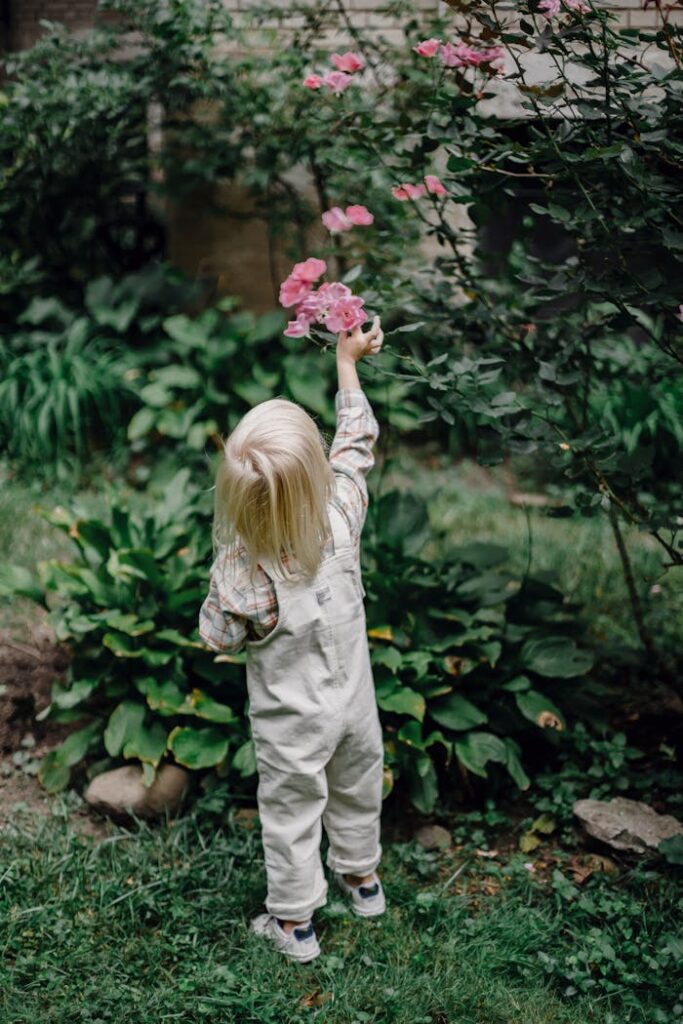 Back view of anonymous little child with blond hair in stylish clothes picking roses from shrub while resting in green park